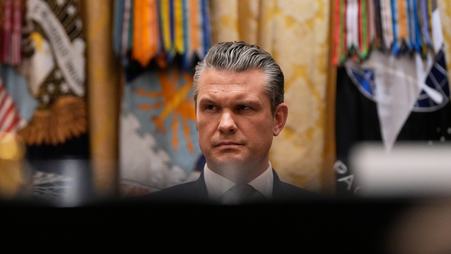 Defense Secretary Pete Hegseth is pictured in front of different flags, partly obscured by shadows.
