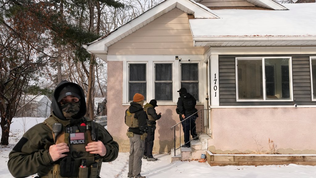 Four officers stand in daylight outside a pink house with snow on the ground. One officer appears in the foreground, wearing a mask, while the other three are gathered in the background