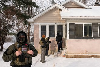 Four officers stand in daylight outside a pink house with snow on the ground. One officer appears in the foreground, wearing a mask, while the other three are gathered in the background