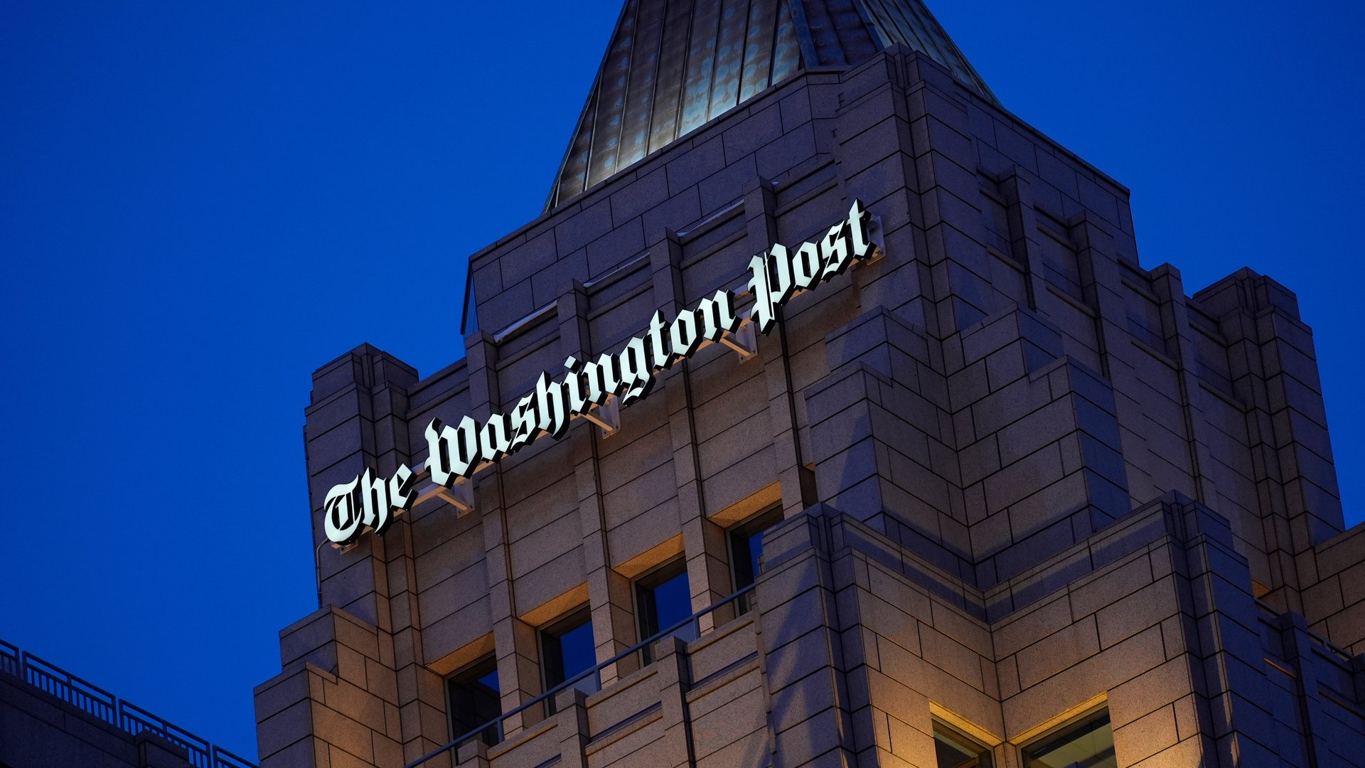 The facade of the Washington Post building shown at dusk, with the words “The Washington Post” lit in white.