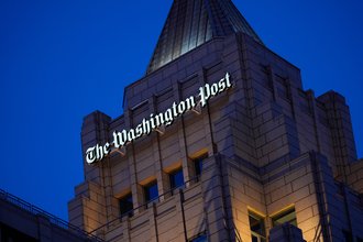 The facade of the Washington Post building shown at dusk, with the words “The Washington Post” lit in white.