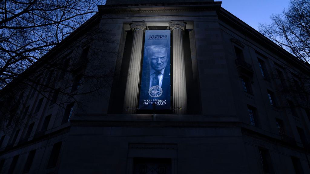 A large banner hanging between the columns of a neoclassical building in Washington, D.C., lit up at dusk, with a portrait of President Donald Trump.