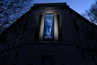A large banner hanging between the columns of a neoclassical building in Washington, D.C., lit up at dusk, with a portrait of President Donald Trump.