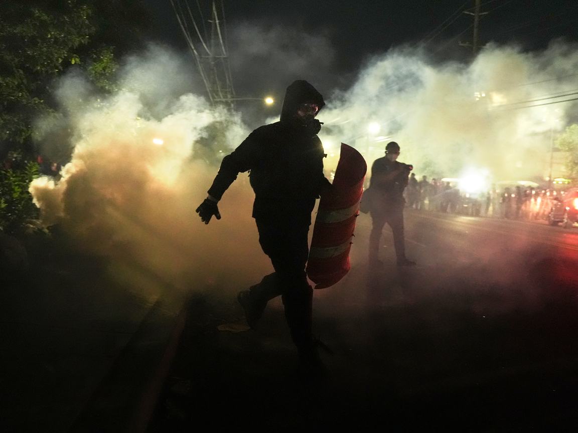 Two figures stand in front of clouds of tear gas; a crowd of protesters is in the background