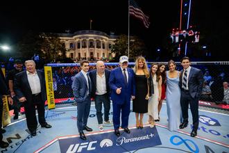 President Donald Trump gives a thumbs up while standing on the Paramount+ logo in a UFC octagon in front of the White House, surrounded by Melania Trump, Dana White, and other guests.