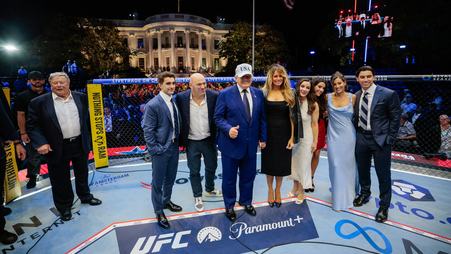 President Donald Trump gives a thumbs up while standing on the Paramount+ logo in a UFC octagon in front of the White House, surrounded by Melania Trump, Dana White, and other guests.