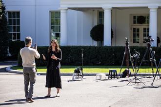 A female news reporter in a black dress stands, with the White House and several TV cameras behind her, speaking toward a man holding up a phone as if to record her.