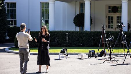 A female news reporter in a black dress stands, with the White House and several TV cameras behind her, speaking toward a man holding up a phone as if to record her.