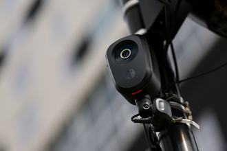 A camera lens in a black, oblong case attached to a pole, shown in front of a blurred background.