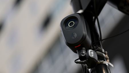 A camera lens in a black, oblong case attached to a pole, shown in front of a blurred background.