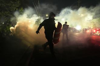 A person in a coat and a gas mask holding half of a traffic cone, surrounded by clouds of tear gas outside the Delaney Hall detention center in Newark, New Jersey