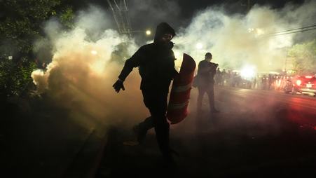 A person in a coat and a gas mask holding half of a traffic cone, surrounded by clouds of tear gas outside the Delaney Hall detention center in Newark, New Jersey