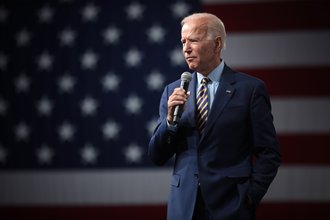 President Biden stands in the foreground wearing a blue suit with a stripped tie and holding a microphone. The American flag is in the background.