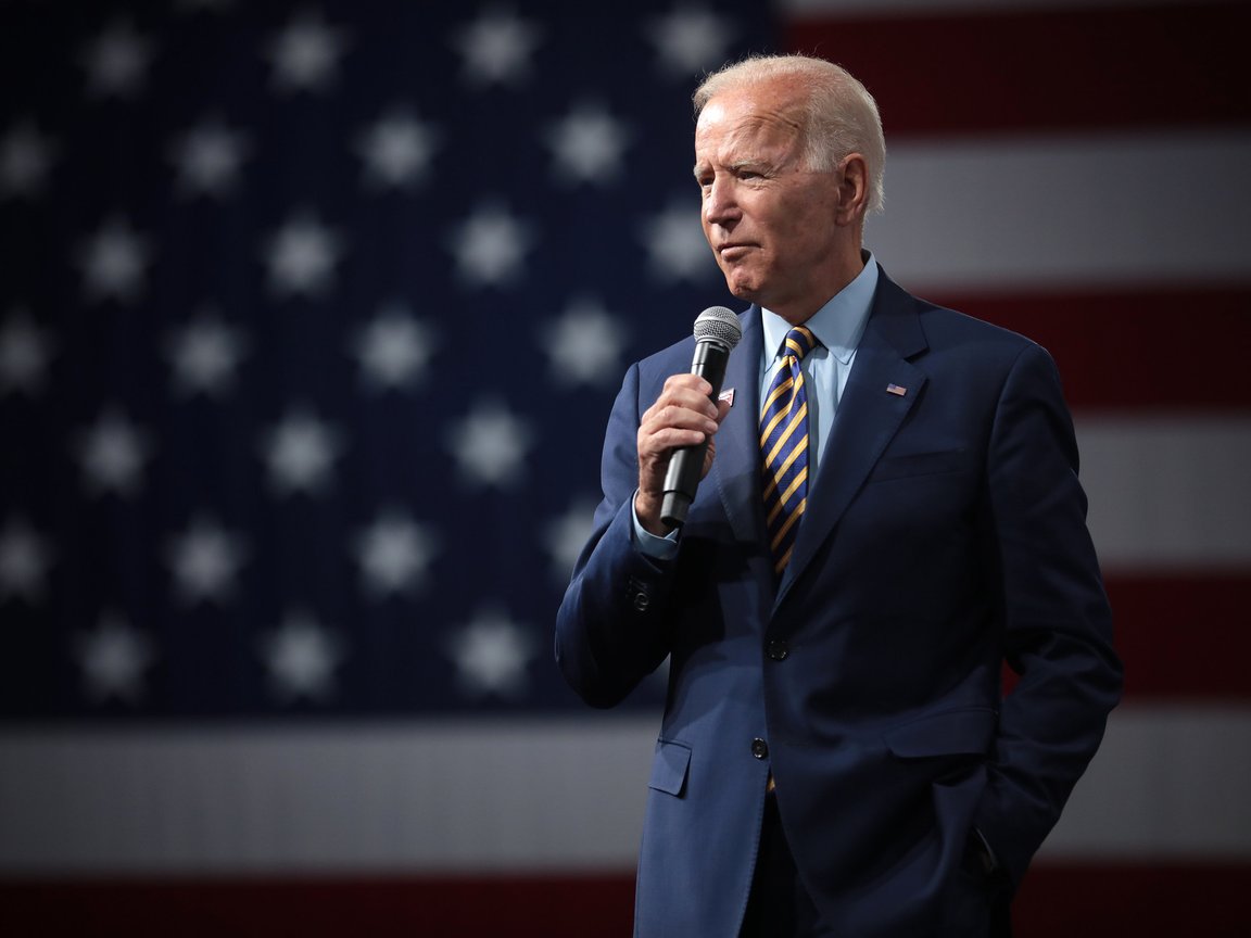 President Biden stands in the foreground wearing a blue suit with a stripped tie and holding a microphone. The American flag is in the background.