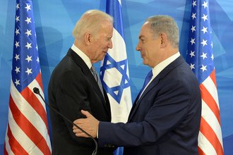 President Biden and President Netanyahu stand facing each other, with Netanyahu's hand on Biden's harm, in front of two American flags and one Israeli flag.