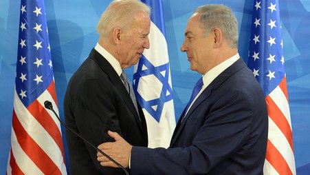 President Biden and President Netanyahu stand facing each other, with Netanyahu's hand on Biden's harm, in front of two American flags and one Israeli flag.