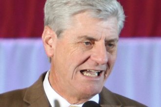 Former Mississippi Gov. Phil Bryant, wearing a brown suit and yellow tie, speaks into a microphone while standing in front of an American flag