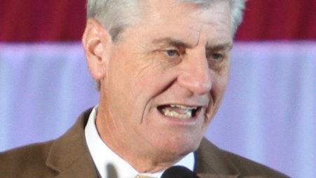 Former Mississippi Gov. Phil Bryant, wearing a brown suit and yellow tie, speaks into a microphone while standing in front of an American flag
