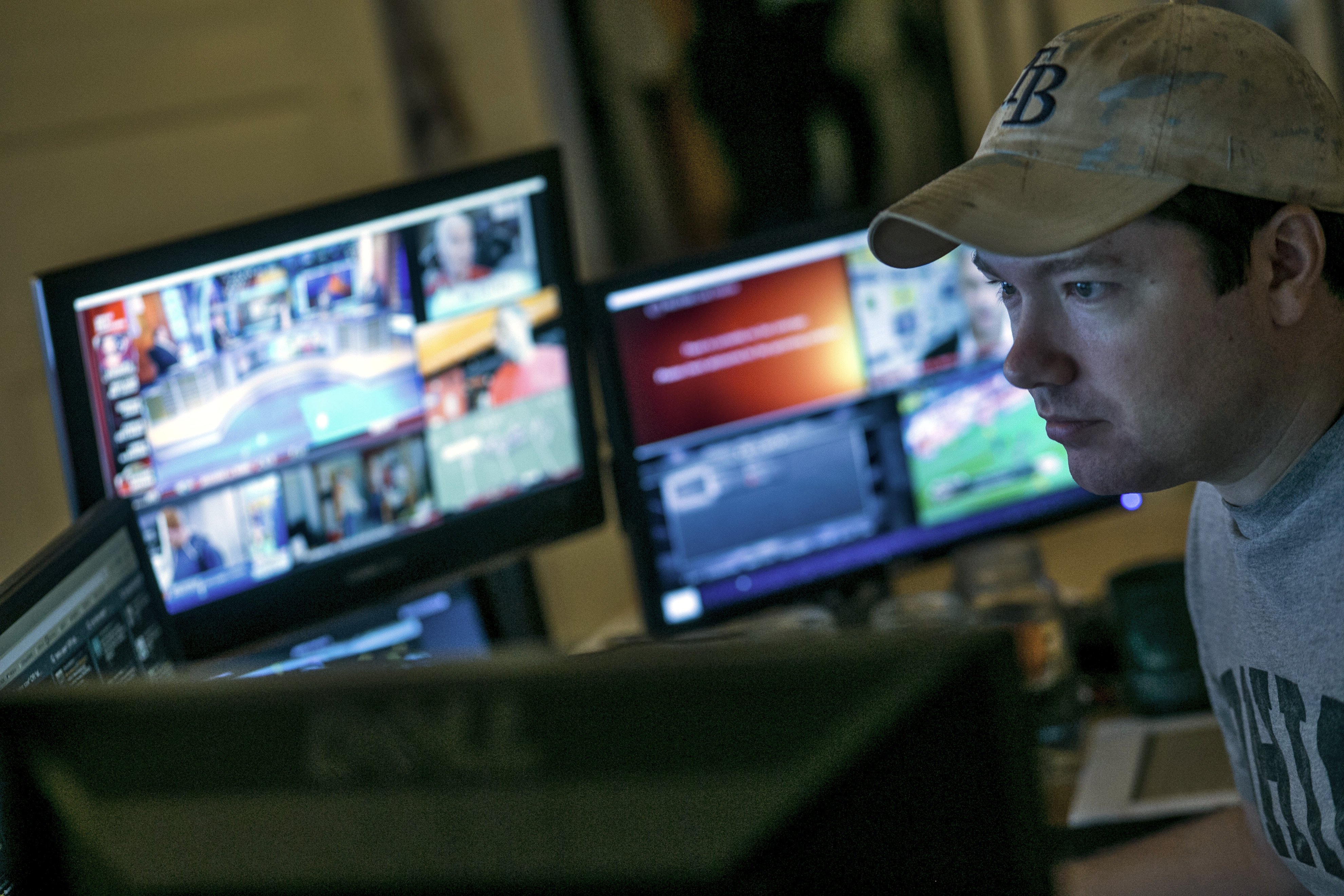 Journalist Tim Burke, wearing a baseball cap, sits in front of computers with blurred images