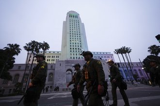 Four officers wearing uniforms and riot hats with face shields walk down the street at dusk, in front of a skyscraper and palm trees.