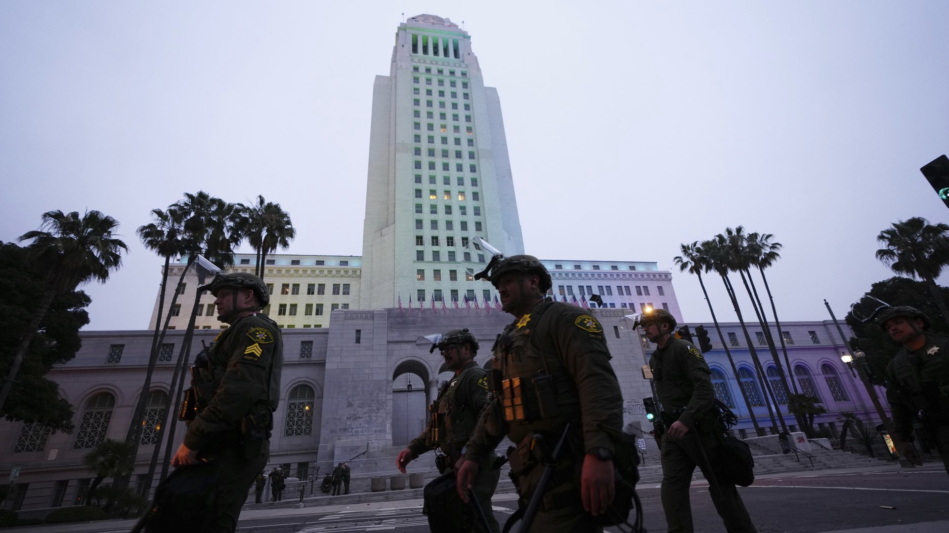 Four officers wearing uniforms and riot hats with face shields walk down the street at dusk, in front of a skyscraper and palm trees.