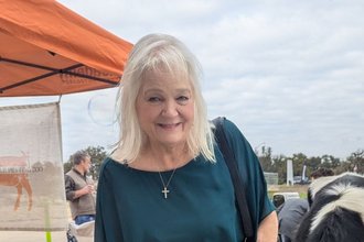 Carol Hemphill, smiling and wearing a blue shirt and cross necklace, stands outside.