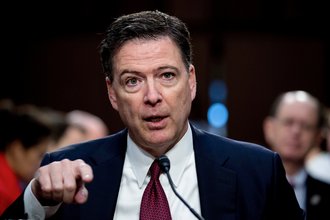 James Comey, pictured from the chest up, speaks into a microphone and points forward while wearing a dark suit and red tie.