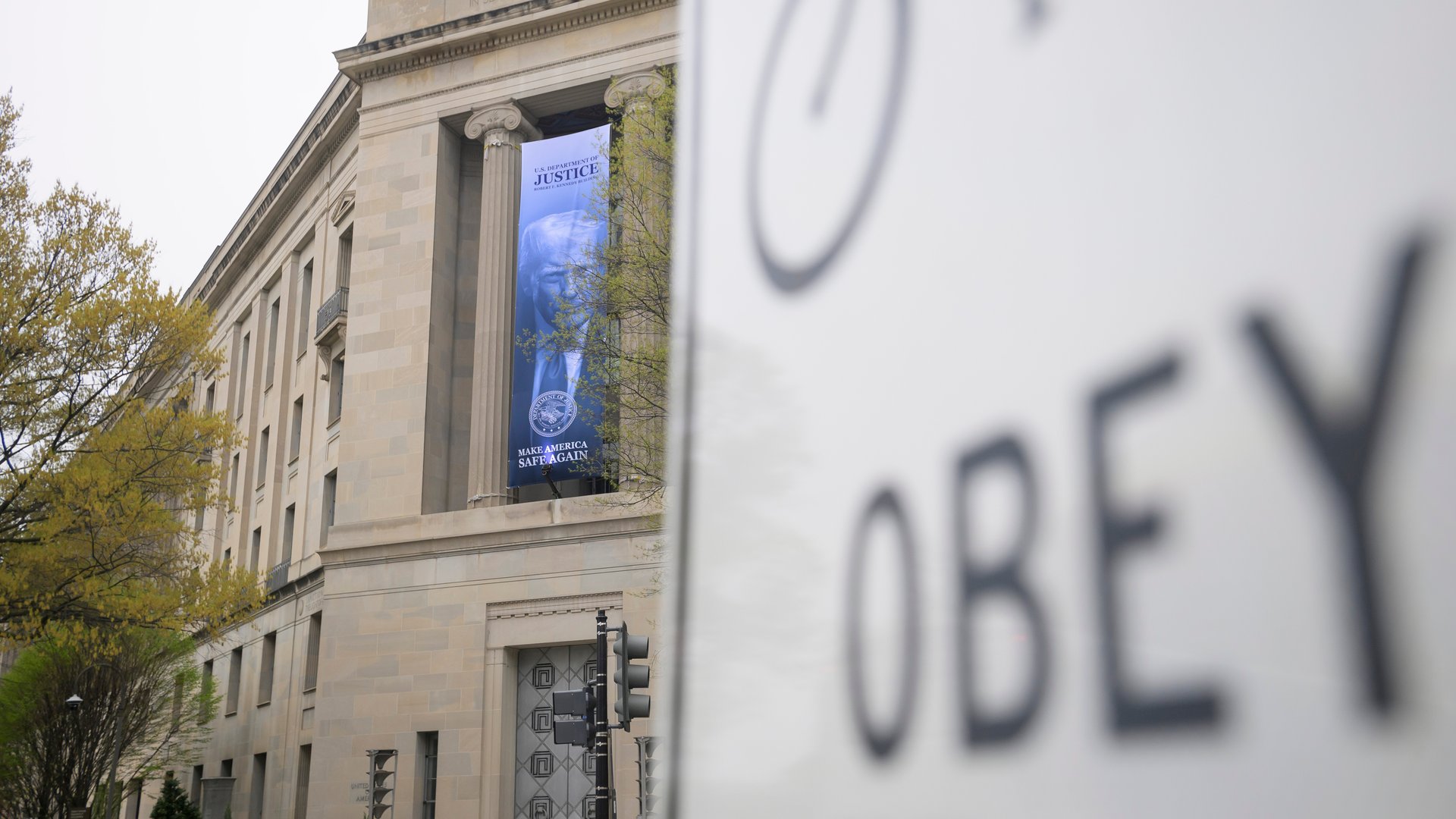 A blue banner showing President Donald Trump’s face hangs from the Department of Justice in the background, and a traffic sign showing the word “OBEY” appears, blurred in the foreground on the right.