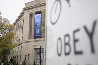 A blue banner showing President Donald Trump’s face hangs from the Department of Justice in the background, and a traffic sign showing the word “OBEY” appears, blurred in the foreground on the right.
