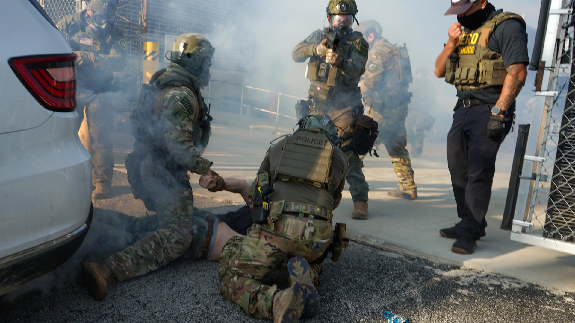 The image shows a group of individuals in tactical gear, mostly obscured by smoke or fog, in what appears to be a training or arrest scenario. Two figures in camouflage and tactical vests with "POLICE" written on them are on the ground restraining a third person who is lying face down. The restraining figures are wearing helmets, and masks or respirators. Another person in similar gear is standing and aiming a rifle. To the right, a person in a black polo shirt, black pants, and a baseball cap is standing and adjusting a mask. A white vehicle is visible on the left. The overall scene is hazy with smoke.