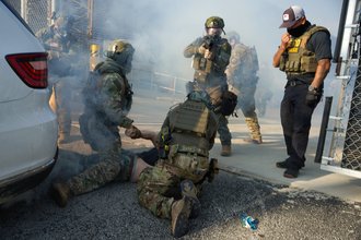 The image shows a group of individuals in tactical gear, mostly obscured by smoke or fog, in what appears to be a training or arrest scenario. Two figures in camouflage and tactical vests with "POLICE" written on them are on the ground restraining a third person who is lying face down. The restraining figures are wearing helmets, and masks or respirators. Another person in similar gear is standing and aiming a rifle. To the right, a person in a black polo shirt, black pants, and a baseball cap is standing and adjusting a mask. A white vehicle is visible on the left. The overall scene is hazy with smoke.