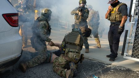 The image shows a group of individuals in tactical gear, mostly obscured by smoke or fog, in what appears to be a training or arrest scenario. Two figures in camouflage and tactical vests with "POLICE" written on them are on the ground restraining a third