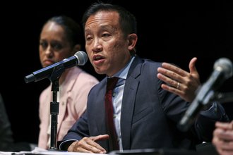 David Chiu sits at a table speaking into a microphone, wearing a grey suit and red tie, and holding up his left hand.