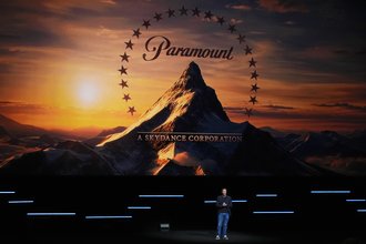 David Ellison, wearing a dark blazer and jeans, speaks on a stage in front of a large Paramount logo.