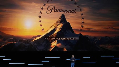 David Ellison, wearing a dark blazer and jeans, speaks on a stage in front of a large Paramount logo.