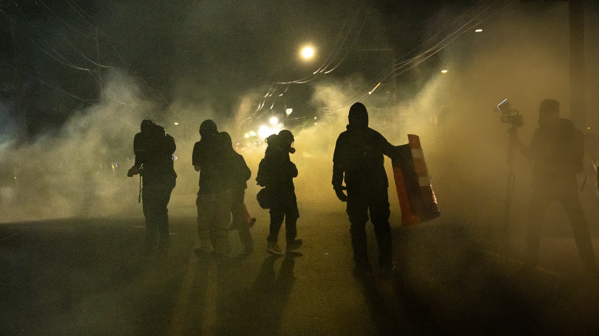 Figures of several people stand on a street at night, surrounded by a cloud of gas. On the right, a person stands behind a camera wearing a face covering.