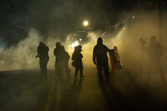 Figures of several people stand on a street at night, surrounded by a cloud of gas. On the right, a person stands behind a camera wearing a face covering.