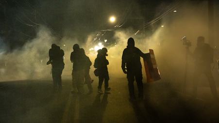 Figures of several people stand on a street at night, surrounded by a cloud of gas. On the right, a person stands behind a camera wearing a face covering.