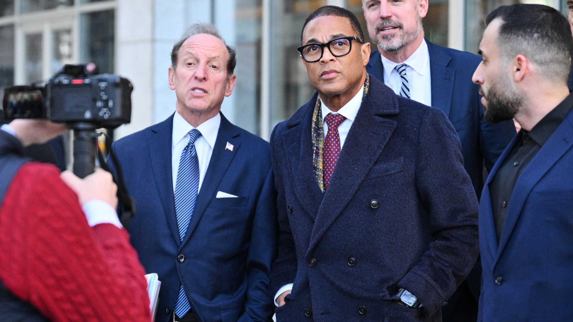 Don Lemon stands outside facing a camera during daytime. Three men in suits stand around him.