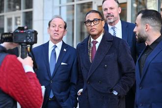 Don Lemon stands outside facing a camera during daytime. Three men in suits stand around him.