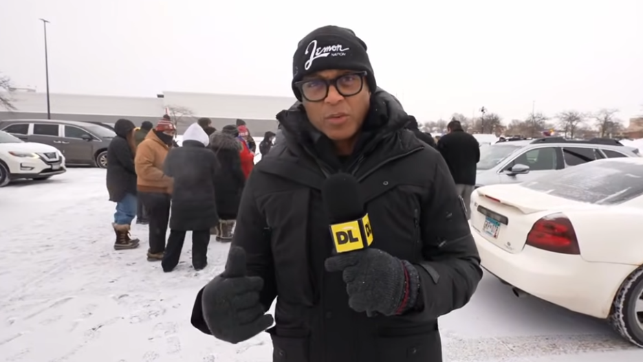 Journalist Don Lemon wearing a black coat and beanie stands with a microphone in front of a crowd of people and cars.