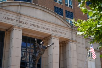 The facade of the Albert V. Bryan United States Courthouse shown in daylight, including a statue of Lady Justice and an American flag.