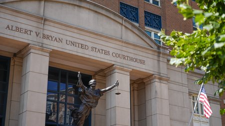 The facade of the Albert V. Bryan United States Courthouse shown in daylight, including a statue of Lady Justice and an American flag.