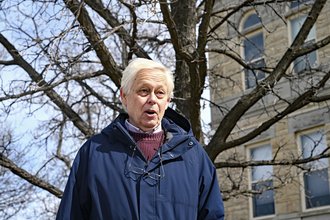Eric Meyer, wearing a blue jacket over a red sweater, with glasses hanging around his neck, stands and speaks in front of a building and a tree.