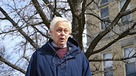 Eric Meyer, wearing a blue jacket over a red sweater, with glasses hanging around his neck, stands and speaks in front of a building and a tree.