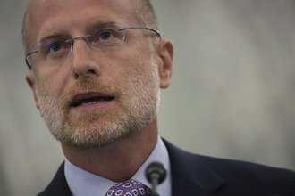 Close-up of Brendan Carr’s face as he speaks into a microphone. Carr is wearing glasses and a dark suit.