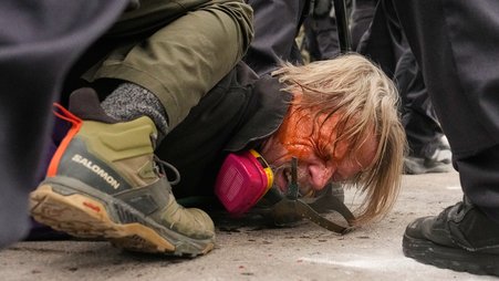 Close-up of the face and torso of a man lying on the ground, grimacing, surrounded by the legs and feet of others, as another person appears to kneel on his back. The man has his eyes squeezed shut and orange liquid running down his face.