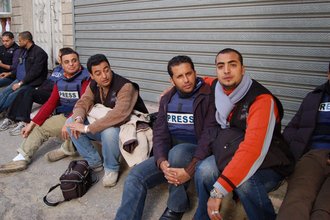A group of male journalists, some wearing blue flak jackets that say PRESS, sit outside a building