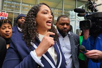 Georgia Fort, shown in profile with her hand raised, wearing a blue blazer and white shirt, speaks while standing outside. In the background are a news camera and microphone, and several people standing around her.