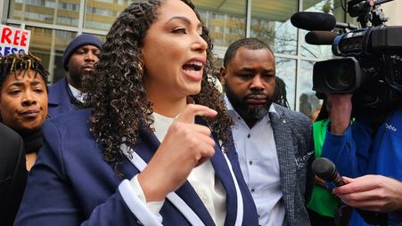 Georgia Fort, shown in profile with her hand raised, wearing a blue blazer and white shirt, speaks while standing outside. In the background are a news camera and microphone, and several people standing around her.